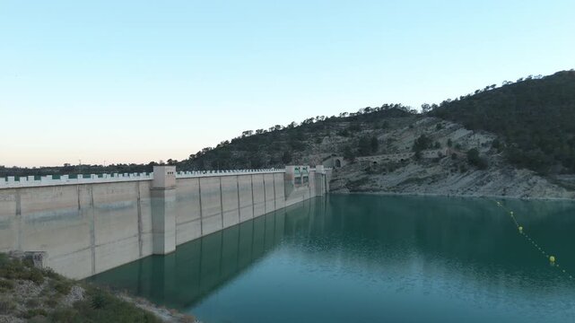 Aerial camera rising from a low angle showing the massive wall of a concrete dam reflecting in the calm green water of a reservoir.