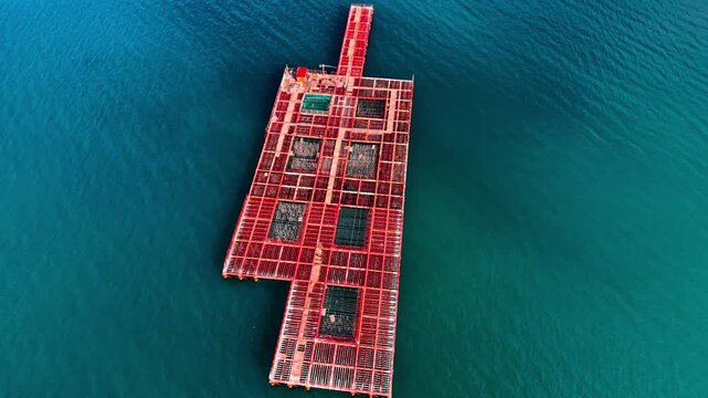 Drone view of red aquaculture platform on teal sea. Elongated fish cage structure with grid frames and net pens surrounded by calm deep green water.
