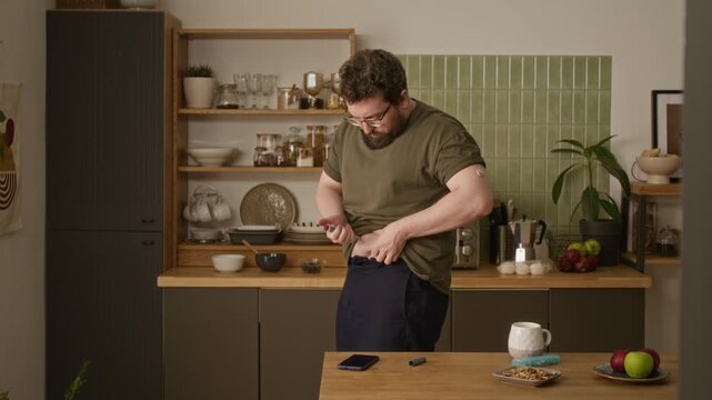 Caucasian middle aged man with diabetes injecting insulin into abdomen with pen injector in kitchen and checking smartphone. Clip ideal for healthcare campaigns and diabetes awareness videos