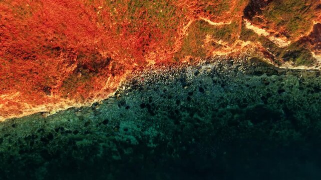 Overhead view of rocky red cliff meeting clear sea. Top down drone perspective of vivid rust red eroded rock surface with scattered boulders transitioning to dark teal transparent water.