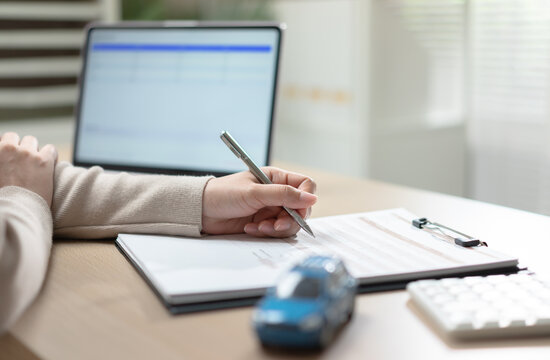 Person signing a vehicle financing document at a desk with a laptop and miniature car, representing auto loan approval, insurance application financial planning, contract agreement, and car ownership.