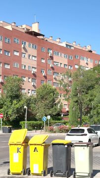 Vertical 4K footage of yellow and gray recycling bins on a sunny Madrid street, with red brick apartment buildings, parked cars, and clear blue sky. No people visible. ES: V&iacute;deo vertical 4K de contene