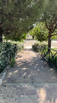 Vertical 4K footage of a shaded brick walkway flanked by classic green metal railings and lush trees in a sunny urban park in Spain. No people visible.
