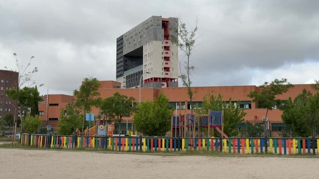 Madrid, Spain, June 9-2026: Urban school playground with colorful fence, play equipment, trees, and educational buildings. Modern high-rise residential architecture visible in the background under an 