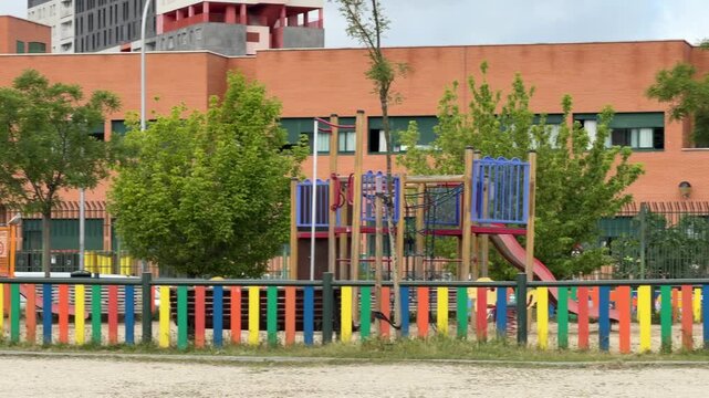 Madrid, Spain, June 9-2026: View of a school playground with colorful fence, play equipment, trees, and modern educational buildings in an urban environment. Exterior scene on a cloudy day.