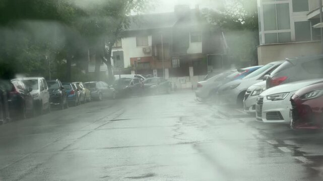 View through a rain-covered windshield during heavy rainfall in a city street. Water droplets blur parked cars, buildings, and the urban environment, creating an atmospheric weather scene.