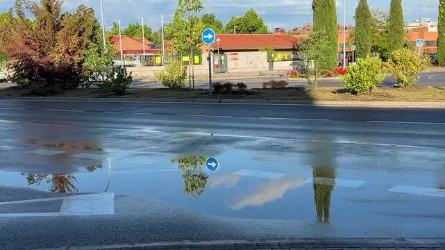 Madrid, Spain, June 9-2026: Urban street after rainfall with large puddle reflecting a passing vehicle, trees, sky, and surrounding buildings. Rainwater reflections and everyday city traffic in an out
