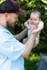 Obraz premium Happy bearded father wearing a baseball cap holding his newborn baby high up in the air while smiling and looking at him in a sunny forest park