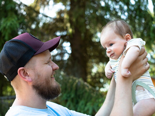 Naklejka premium Happy bearded father wearing a baseball cap holding his newborn baby high up in the air while smiling and looking at him in a sunny forest park