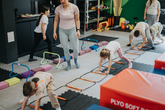 A group of children doing fitness drills with an instructor in an indoor gym. Young kids perform ladder and hurdle exercises during a supervised fitness class.