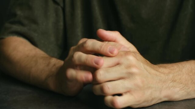 Close up nervous man wringing hands on dark table, showcasing anxiety, stress, and inner turmoil through restless and agitated body language while sitting alone. Anxious man rubbing and wringing hands