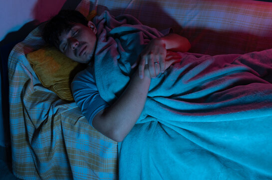 Man resting on a couch at night with a blanket, illuminated by the light of a screen. Rest, comfort, home, and nighttime lifestyle concept