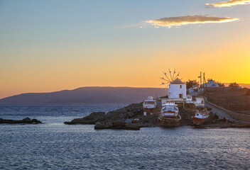 Fototapeta premium Traditional Greek windmill and fishing boats at sunset on a rocky coastline in the Ano Koufonisi island, Greece