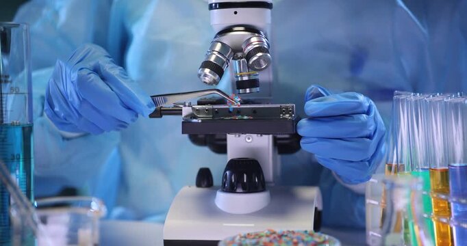 Technician places microplastic particles onto microscope slide in lab setting. Assistant in protective suit analyzes polymer materials in laboratory