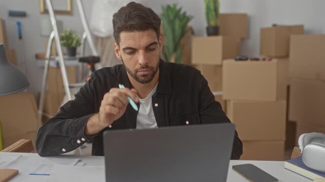Hispanic man with beard holds pen to temple while working on laptop at desk amid moving boxes, lamp and paperwork in studio; pensive planning.