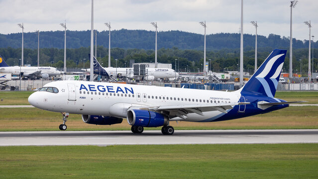 Ein Airbus A320 der Aegean Airlines rollt auf der Startbahn. Im Hintergrund sind weitere Maschinen der Lufthansa am Terminal zu erkennen.