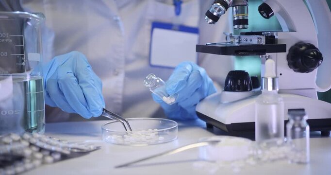 Pharmacist hands fill glass bottle with small pills at table in lab. Medical worker packs experimental supplement by microscope in chemical laboratory
