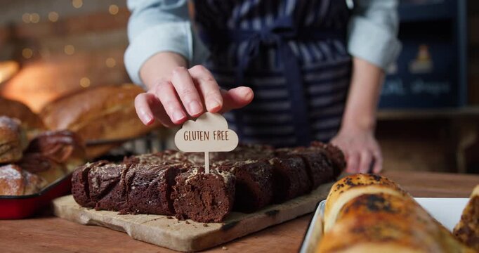Female Sales Assistant In Bakery Putting Gluten Free Label Into Tray Of Brownies