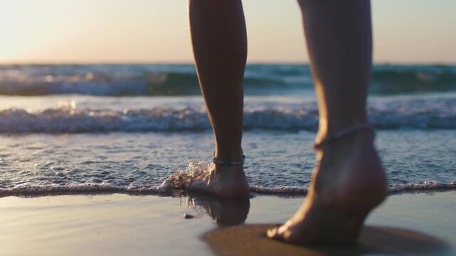 A rear view of a girl's legs and bare feet as she walks along the shoreline on wet sand during golden hour, with gentle waves, warm backlighting and tranquility in the background.