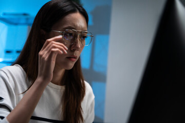 Fototapeta premium Focused Asian woman software engineer adjusting eye glass while coding and looking at computer screen, concentrated female programmer working late on cybersecurity project in office