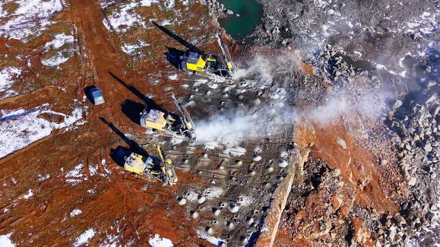 Overhead drone view of drilling rigs at a quarry site, Bulgaria. Top down shot of yellow machines creating dust clouds while boring holes into the rocky ground.