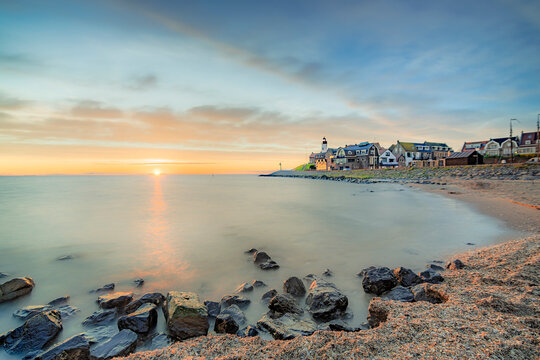 The iconic lighthouse in Urk stands sentinel over a tranquil waterfront as the sun dips below the horizon, casting a warm, golden glow across the calm sea and sky.