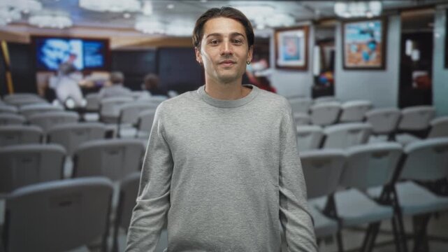 Man shrugging with visible face, wearing a grey sweater, standing before rows of folding chairs and a large display in a building; casual uncertainty.
