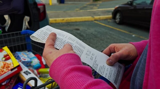 A person checks a shopping list while standing next to a cart full of items in the grocery store parking lot. The list helps in choosing products