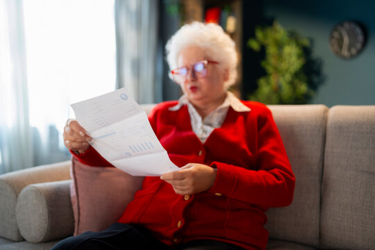 Concerned elderly woman reviewing utility bill and household expenses while sitting on couch at home. Inflation, rising energy costs and retirement finances concept.