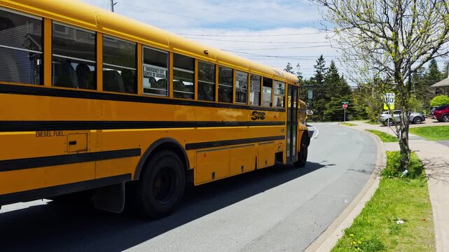 School buses travel on a road in Halifax, Nova Scotia. Bright colors of the buses stand out against the clear sky. Children wait nearby to board the buses