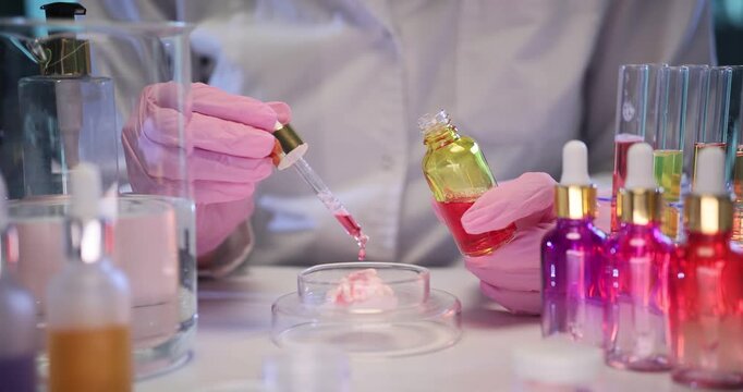 Pharmacist drips essential oil onto cream in glass Petri dish in medical lab. Female dermatologist creates innovative skincare product at workplace