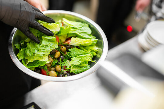 Chef with black gloves preparing a fresh Mediterranean salad with romaine lettuce, olives, and tomatoes in a metal bowl.