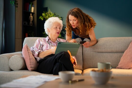 Adult daughter helping elderly mother use tablet and credit card to pay household bills online. Digital banking and financial literacy concept.