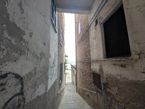 Un vicolo stretto con muri vissuti e gradini in pietra scende verso una zona illuminata dal verde, creando un&rsquo;atmosfera urbana antica e suggestiva ricca di texture e profondit&agrave;