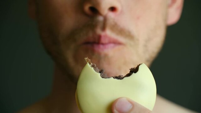 Close up man mouth eating delicious yellow glazed chocolate cookie, showcasing enjoyment and indulgence in sweet treat against simple dark background. Hungry man eating lemon chocolate cookie close up