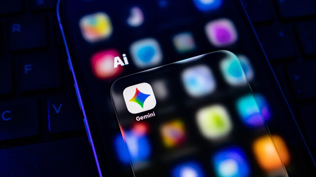Closeup of a smartphone screen displaying the google gemini app icon with a blurred background of app icons and a computer keyboard