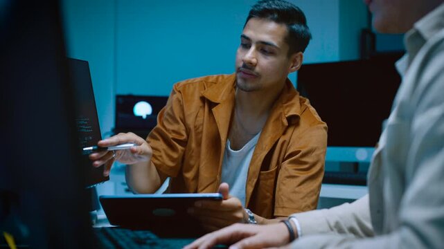 Two happy young male IT specialists smiling and discussing computer software programming code with digital tablet while working together in modern corporate office room