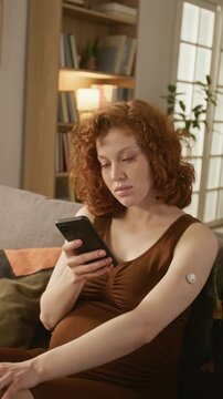 Vertical shot of pregnant Caucasian young adult woman scanning continuous glucose monitor on arm with smartphone. Clip useful for gestational diabetes and maternal healthcare ads