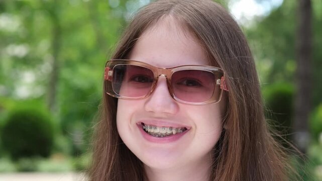 Portrait of caucasian happy teen girl with dental braces outdoors on sunny summer day. Person in sunglasses smiling showing teeth. Happy teenager looking at camera