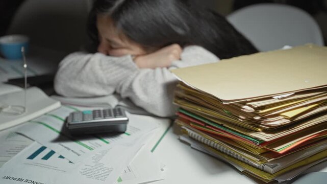 Young chinese woman student rests head on arms over open book, calculator and stacked folders at study desk; exam stress fatigue.
