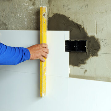 Close up of a bricklayer's hand checking the correct placement of white porcelain stoneware tiles with a level during a complete kitchen renovation in a home.