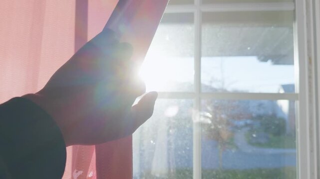 A hand pulls back pink curtains to let in warm light from the sun. The bright rays fill the room as the day begins. The view outside shows a bright morning