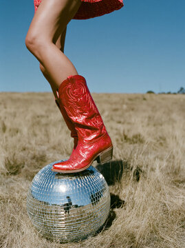 Legs in shiny red cowboy boots step on a mirrored disco ball in a dry field under clear blue sky. Bold western-fashion concept designed for editorial, advertising, lifestyle and social media.