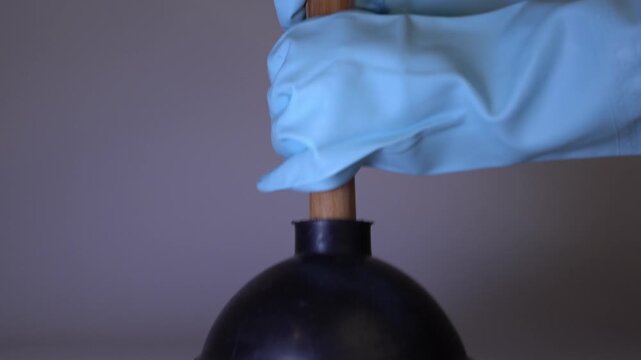 Gloved hand holds a black rubber plunger with a wooden handle, showcasing the tool's details and functionality in a neutral background setting