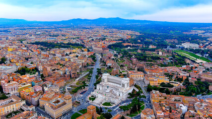 Naklejka premium Aerial drone view of Altare della Patria, the Roman Forum, and the Colosseum in the city center of Rome, Italy, featuring urban streets and distant mountains on a sunny day.