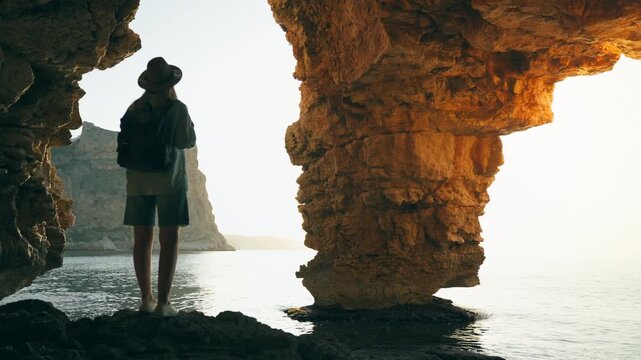 Young woman with backpack standing inside sea cave and looking toward ocean cliffs at sunset, symbolizing self discovery, personal journey and inner freedom.