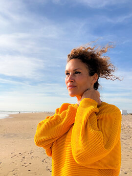 Woman in a yellow sweater standing on a windy beach, portrait of a contemplative middle-aged woman looking toward the horizon in warm sunlight, casual fashion and peaceful coastal scene.