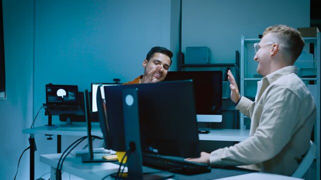 Two young male software developers giving high five celebrating successful coding project completion while working with computers in modern IT office late at night