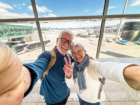 Senior couple taking a joyful airport selfie with backpacks and scarves, posing with peace sign in terminal overlooking airplanes and jet bridge before boarding for travel vacation