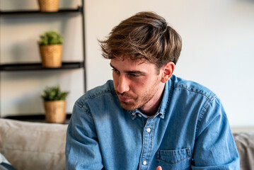 Obraz premium Pensive young man in a denim shirt sitting on a sofa at home, looking down thoughtfully in a casual interior with potted plants — lifestyle portrait conveying contemplation and quiet emotion.